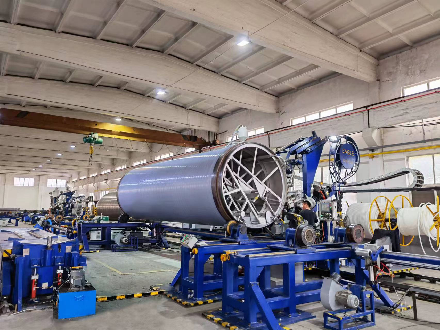 Large spiral-winding pipe machine with a wound HMPP pipe section in a workshop, an operator at the control station